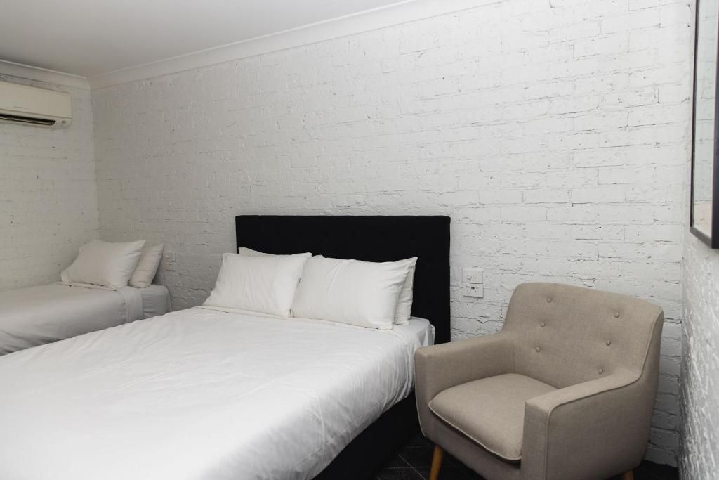 A hotel room with a white brick wall, a black headboard, a single bed, a chair, and a small table.