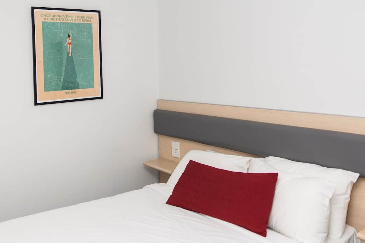 A bedroom with a bed, a nightstand, a red pillow, and a framed poster on the wall.