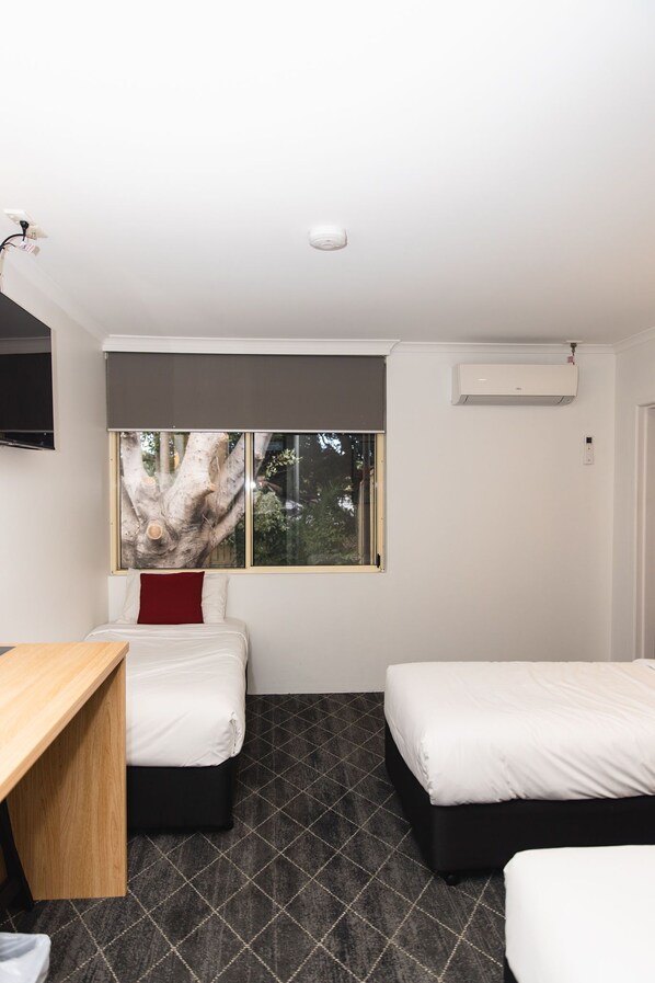 A hotel room with two beds, a desk, a large window with a tree view, and an air conditioning unit.