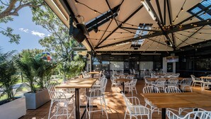 An outdoor dining area with multiple tables and chairs, surrounded by potted plants and a large umbrella providing shade.