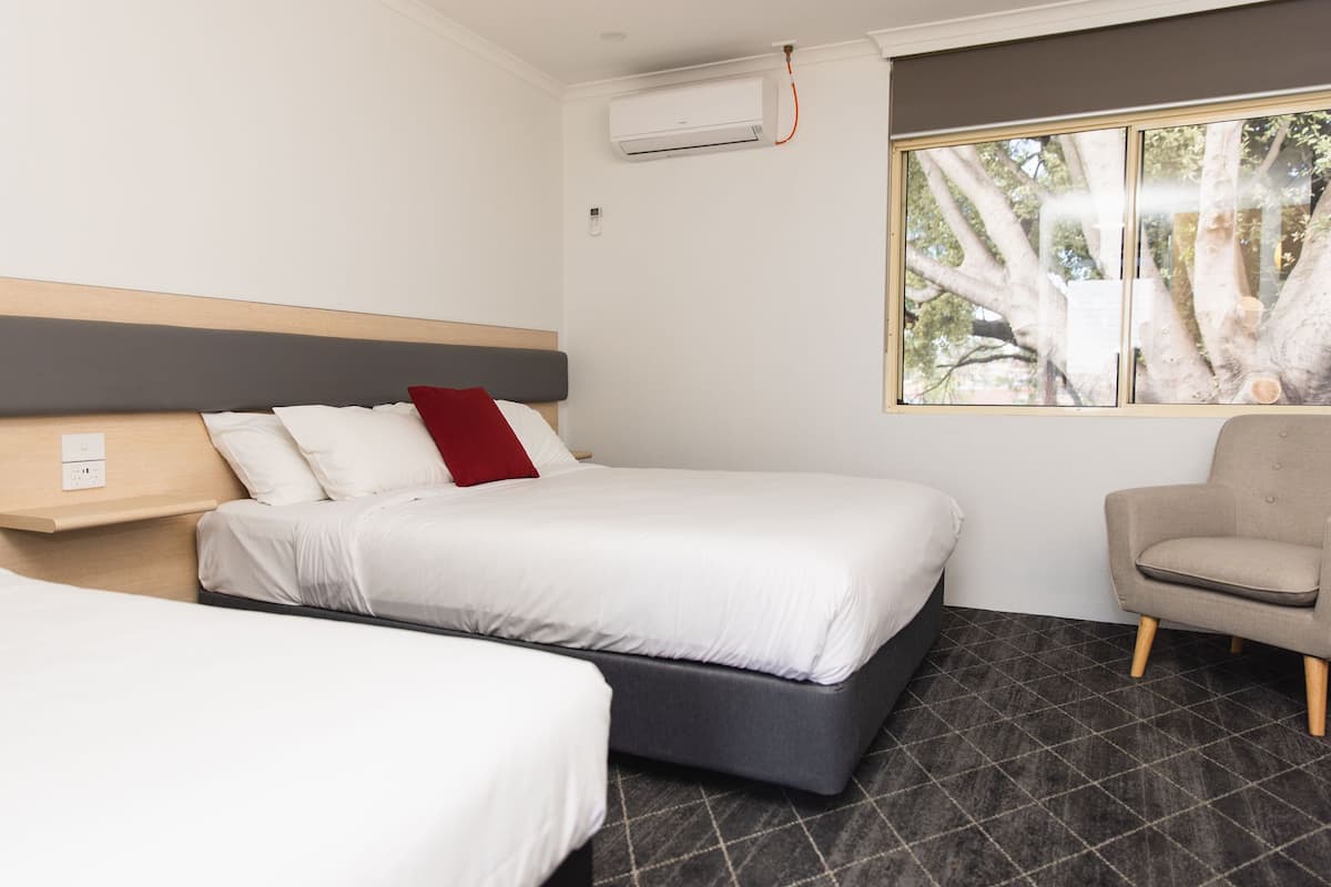 A hotel room with a bed, a chair, a window with a view of trees, and an air conditioning unit.