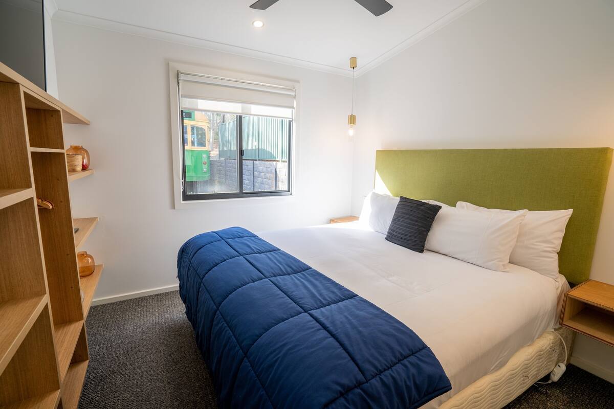 Una habitación de hotel con cama, ventana, estante y mesita de noche.