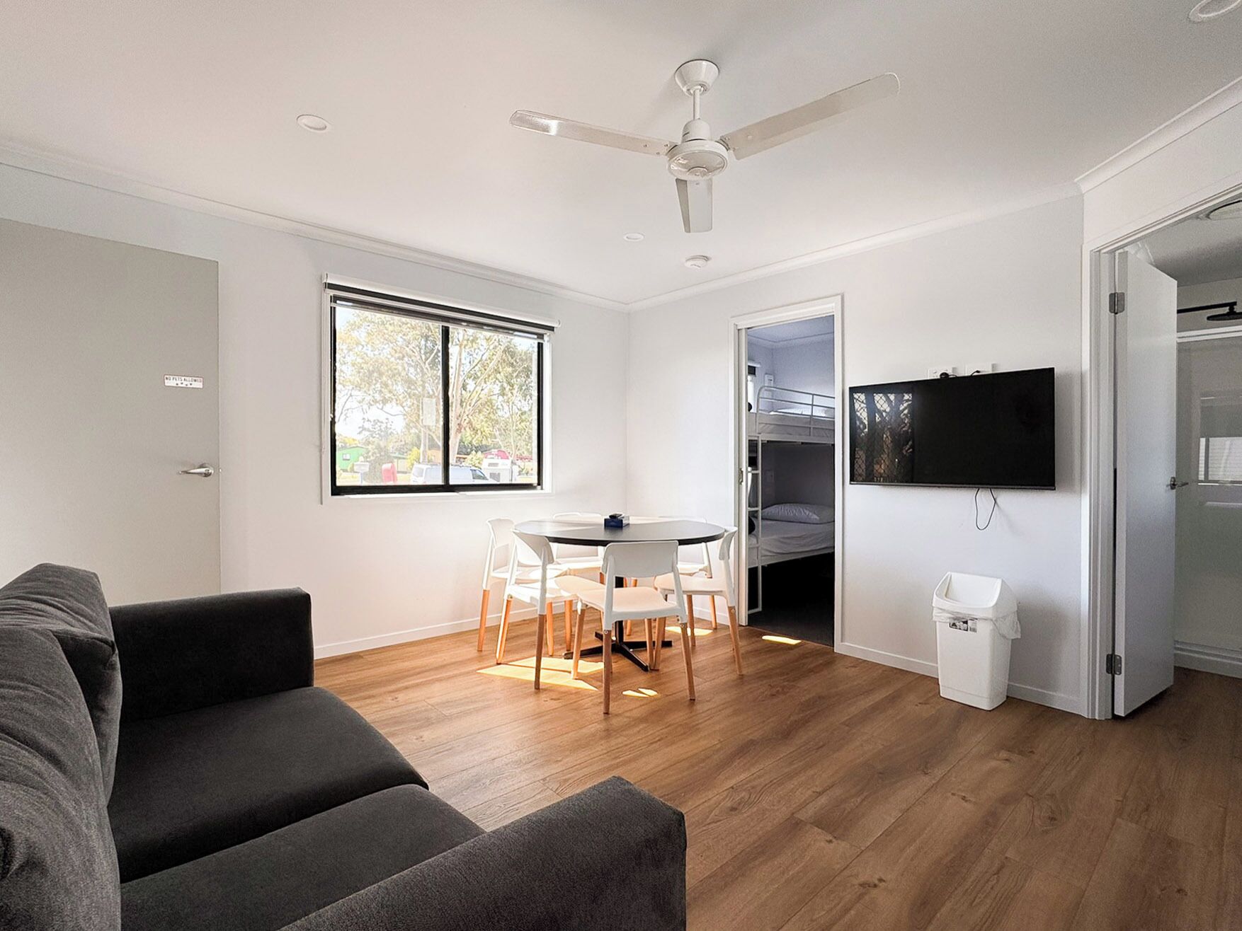 A modern living room with a sofa, a dining table with chairs, a ceiling fan, a TV mounted on the wall, and a window with a view of trees.