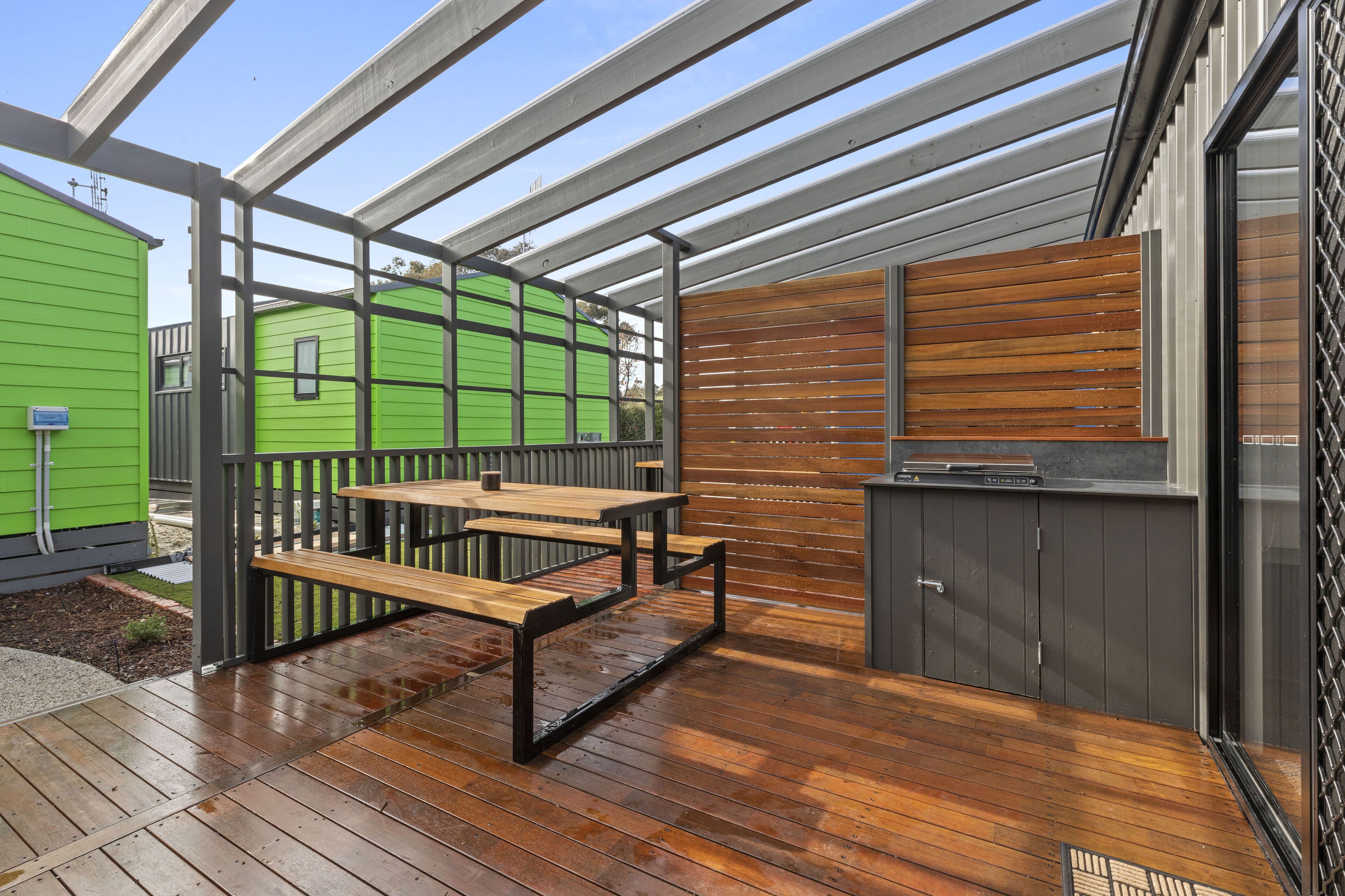 Un patio couvert avec une table et des bancs en bois, un barbecue, et un bâtiment vert en arrière-plan.
