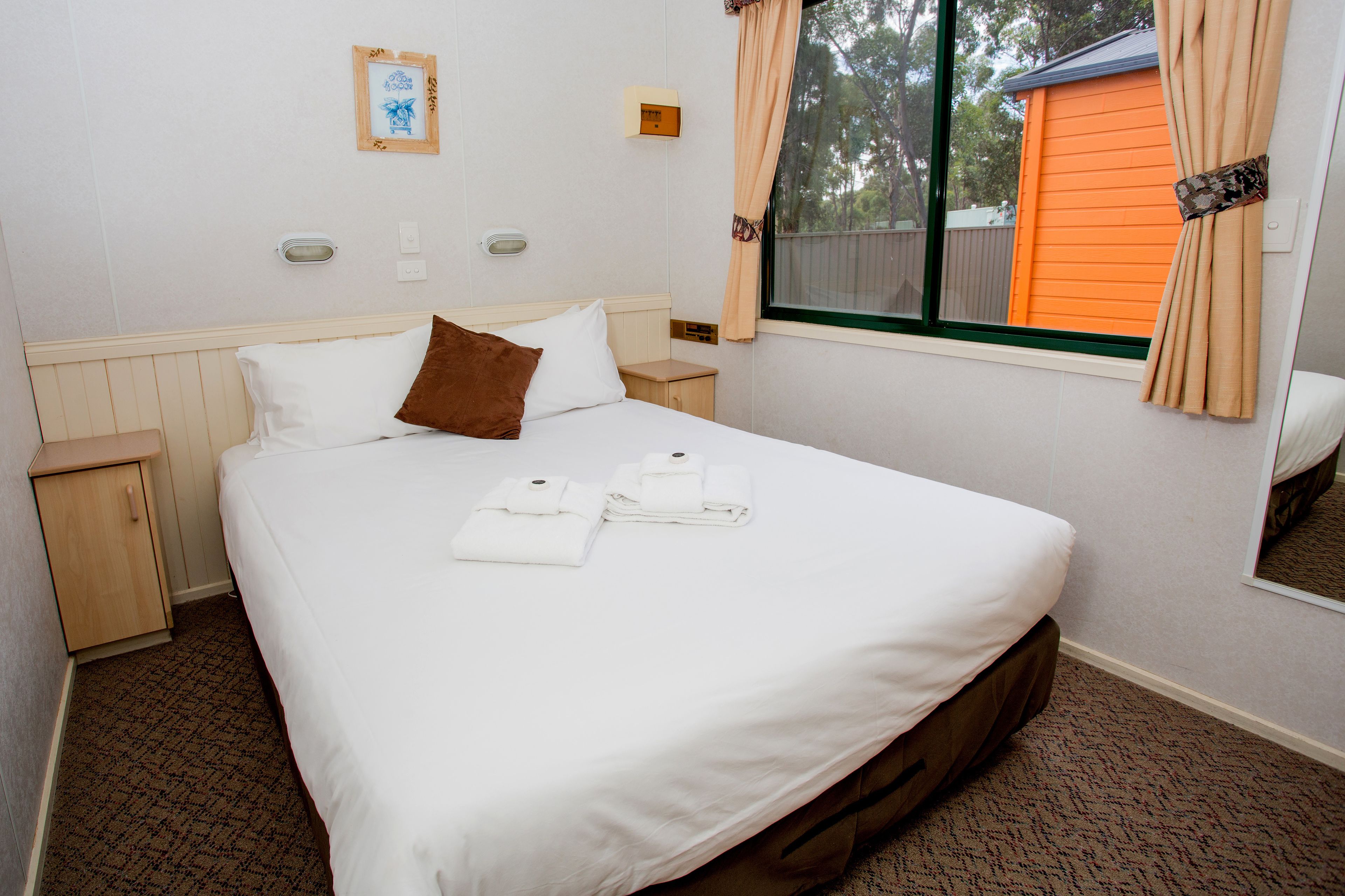 A neatly made bed with white linens and a brown pillow, a wooden nightstand, and a window with curtains.