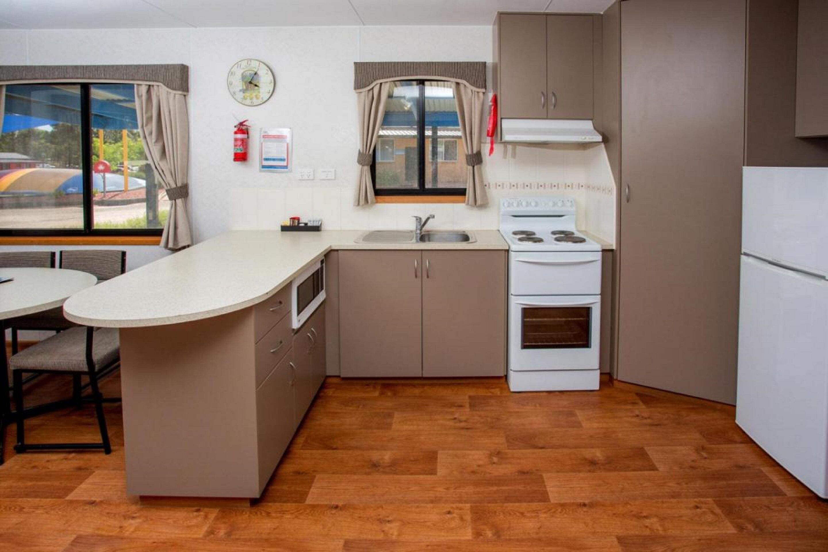 A compact kitchen with a sink, stove, and refrigerator. It features a window with curtains, a clock on the wall, and a fire extinguisher.
