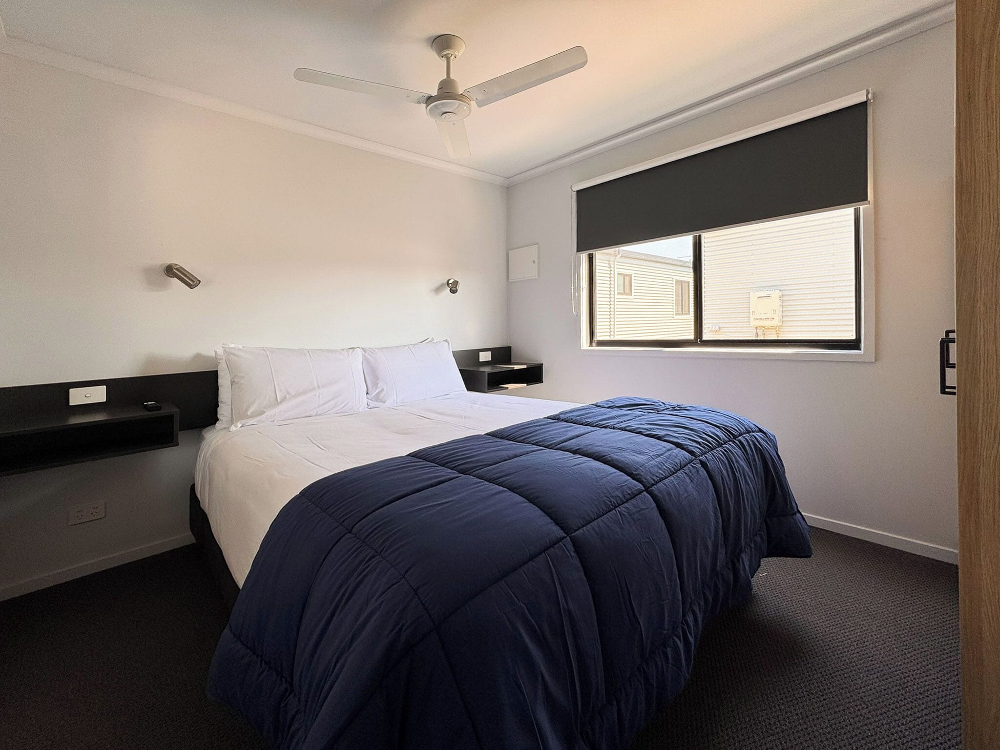 A hotel room with a bed, a ceiling fan, a window with blinds, and a nightstand.