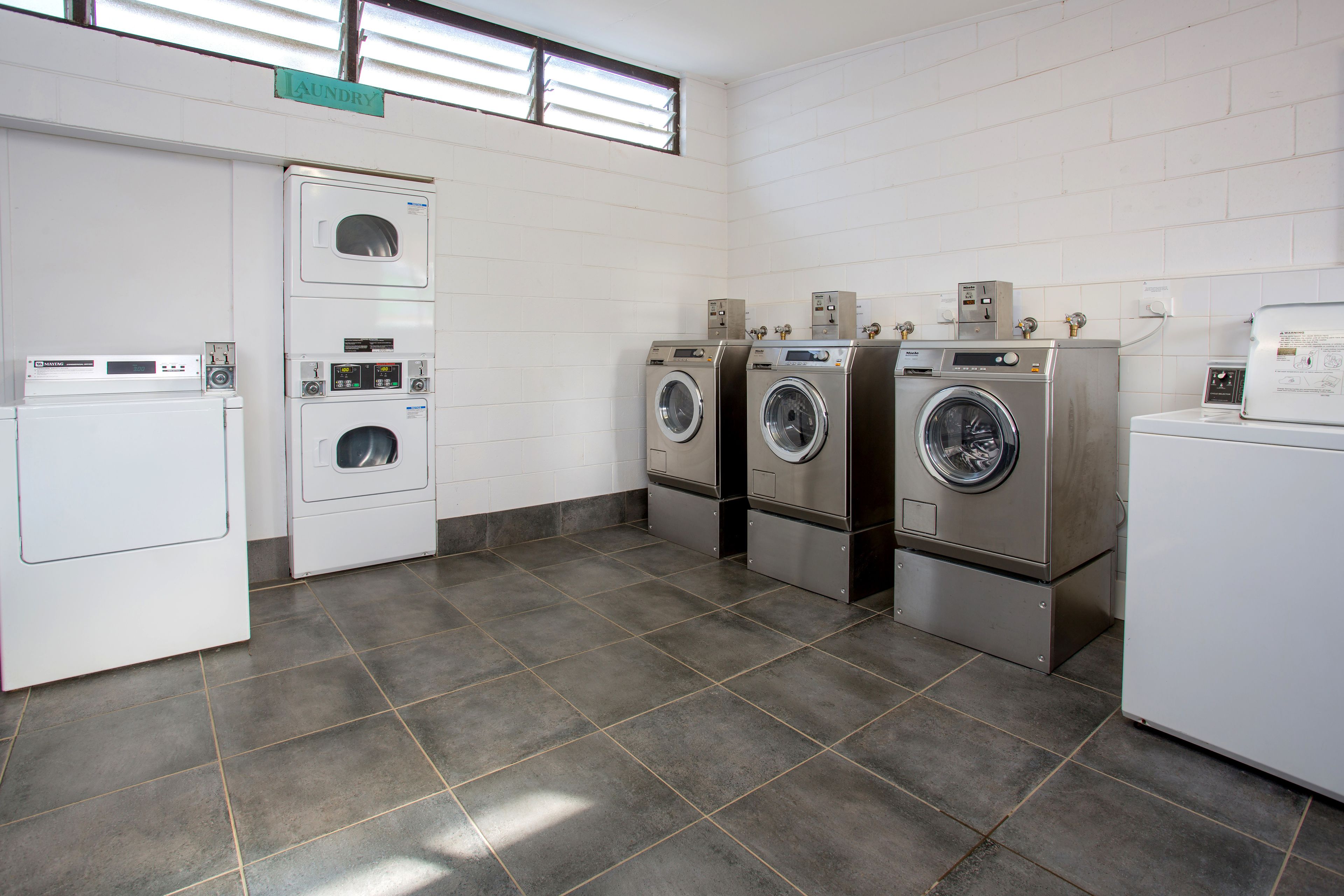 A laundry room with four washing machines and a dryer, white tiled walls, and a tiled floor.