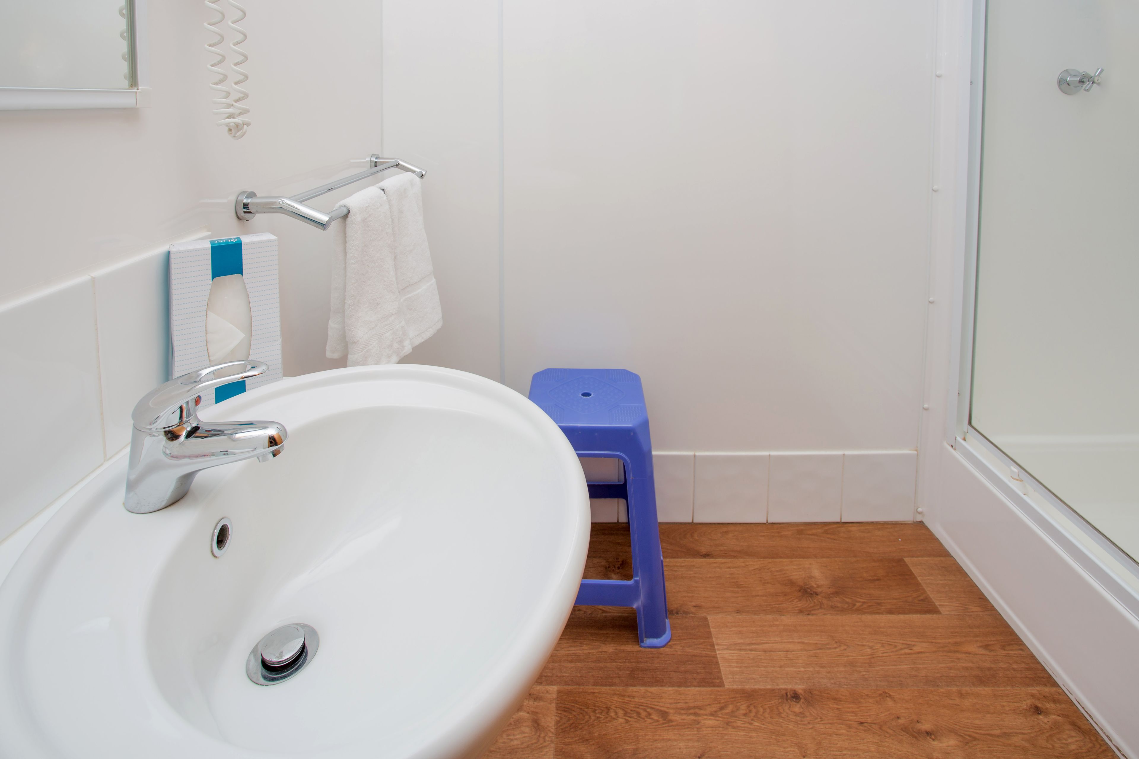 A bathroom with a white sink, a blue stool, and a glass shower door.
