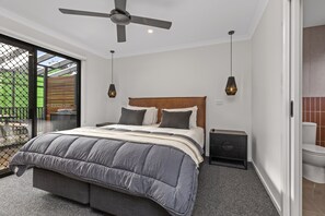 Ein Schlafzimmer mit einem großen Bett, einem Deckenventilator, zwei Pendelleuchten, einem Nachttisch und Blick ins Freie durch eine Schiebetür.
