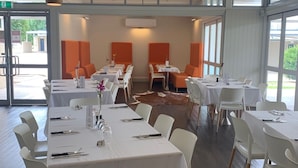 A spacious dining area with white tables and chairs, large windows, and a high ceiling with a central light fixture. - Banana Hotel Motel (Banana)