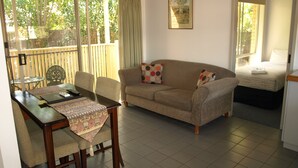 Una sala de estar con un sofá, una mesa de comedor con sillas, una mesita con un camino de mesa decorativo y una cama al fondo.