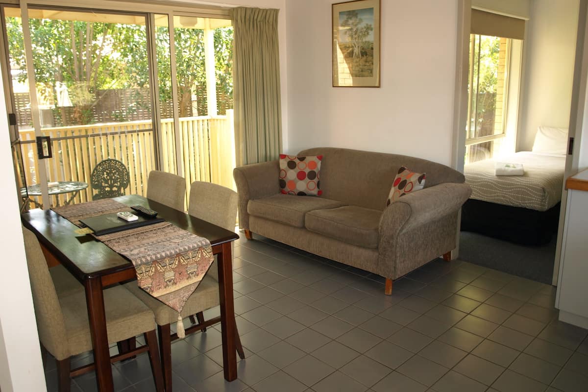 A living room with a sofa, a dining table with chairs, a small table with a decorative table runner, and a bed in the background.