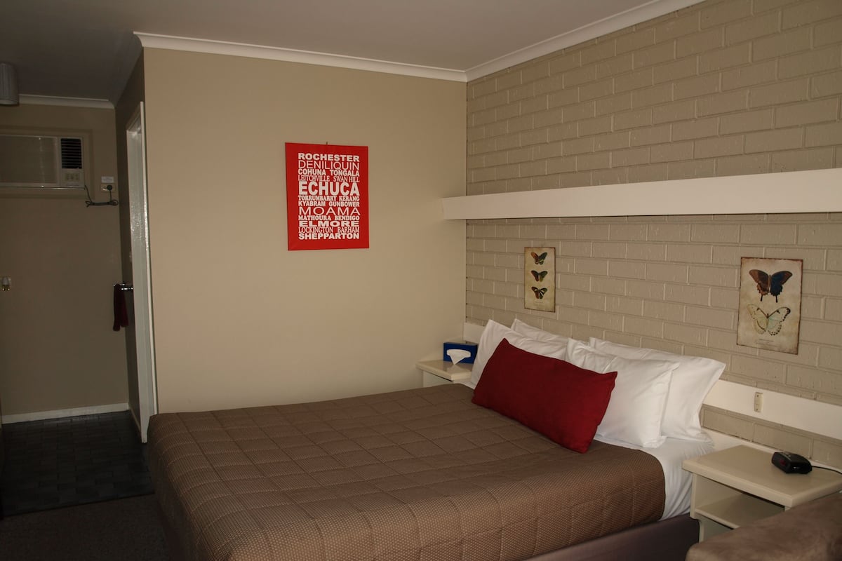 A hotel room with a bed, a nightstand, a red pillow, a wall-mounted picture frame, and a red sign listing local attractions.