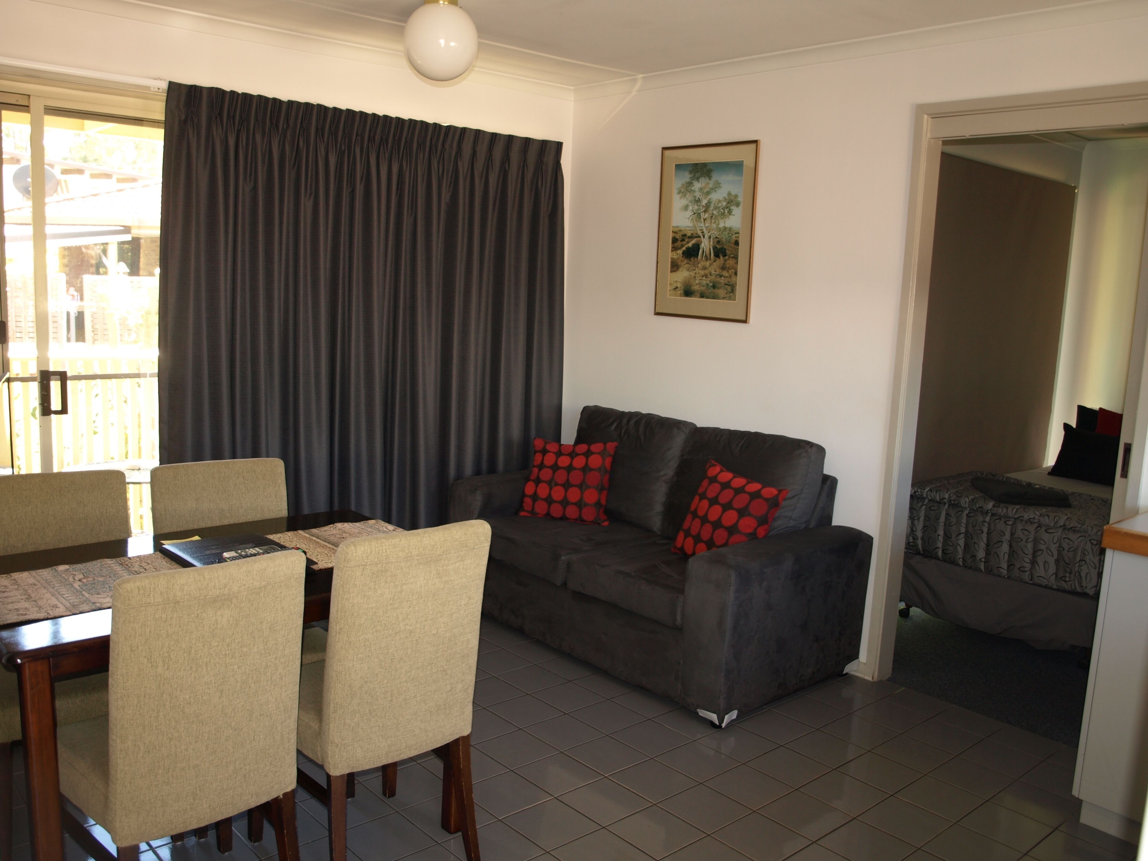 A living room with a dark grey sofa, beige chairs, a dining table with a patterned table runner, and a large window with dark curtains.