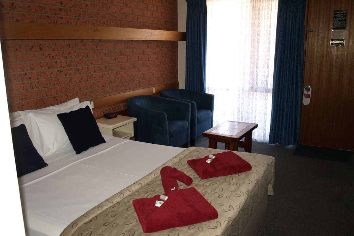 A hotel room with a brick wall, a bed with white and dark blue bedding, two blue armchairs, a small wooden table, and a door with a sign.