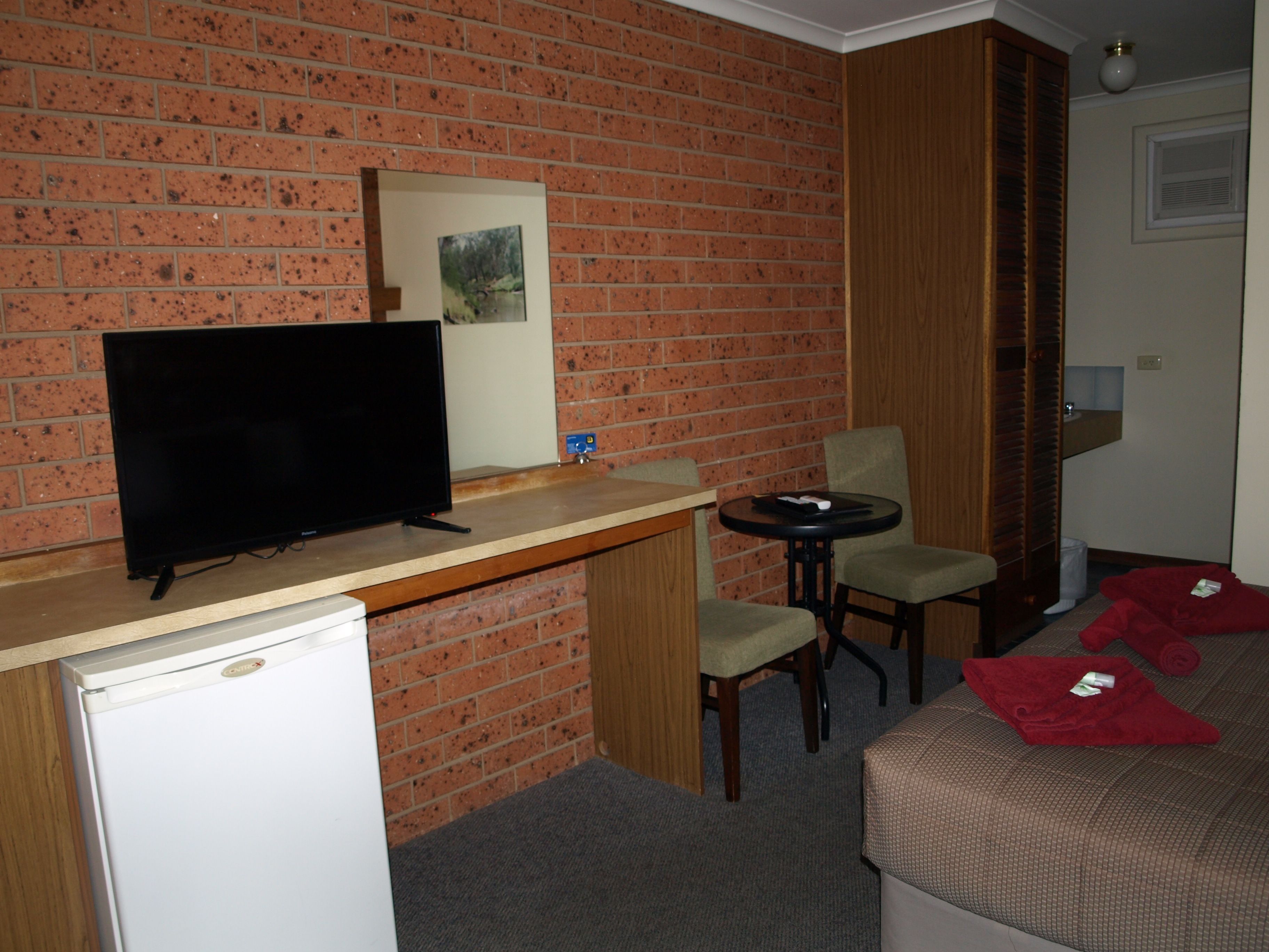 A compact room with a brick wall, a television on a wooden counter, a small table with two chairs, and a bed with a red blanket.
