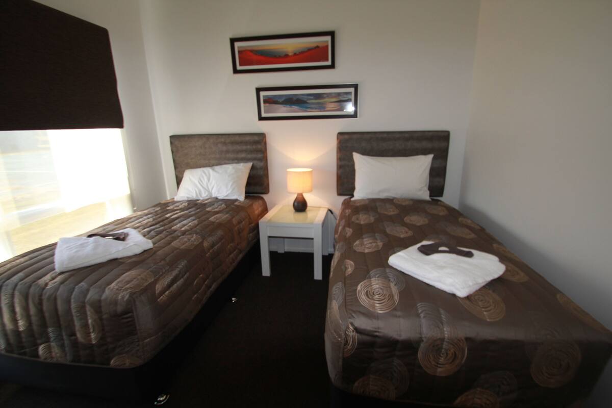 A hotel room with two single beds, a nightstand with a lamp, and two framed pictures on the wall.