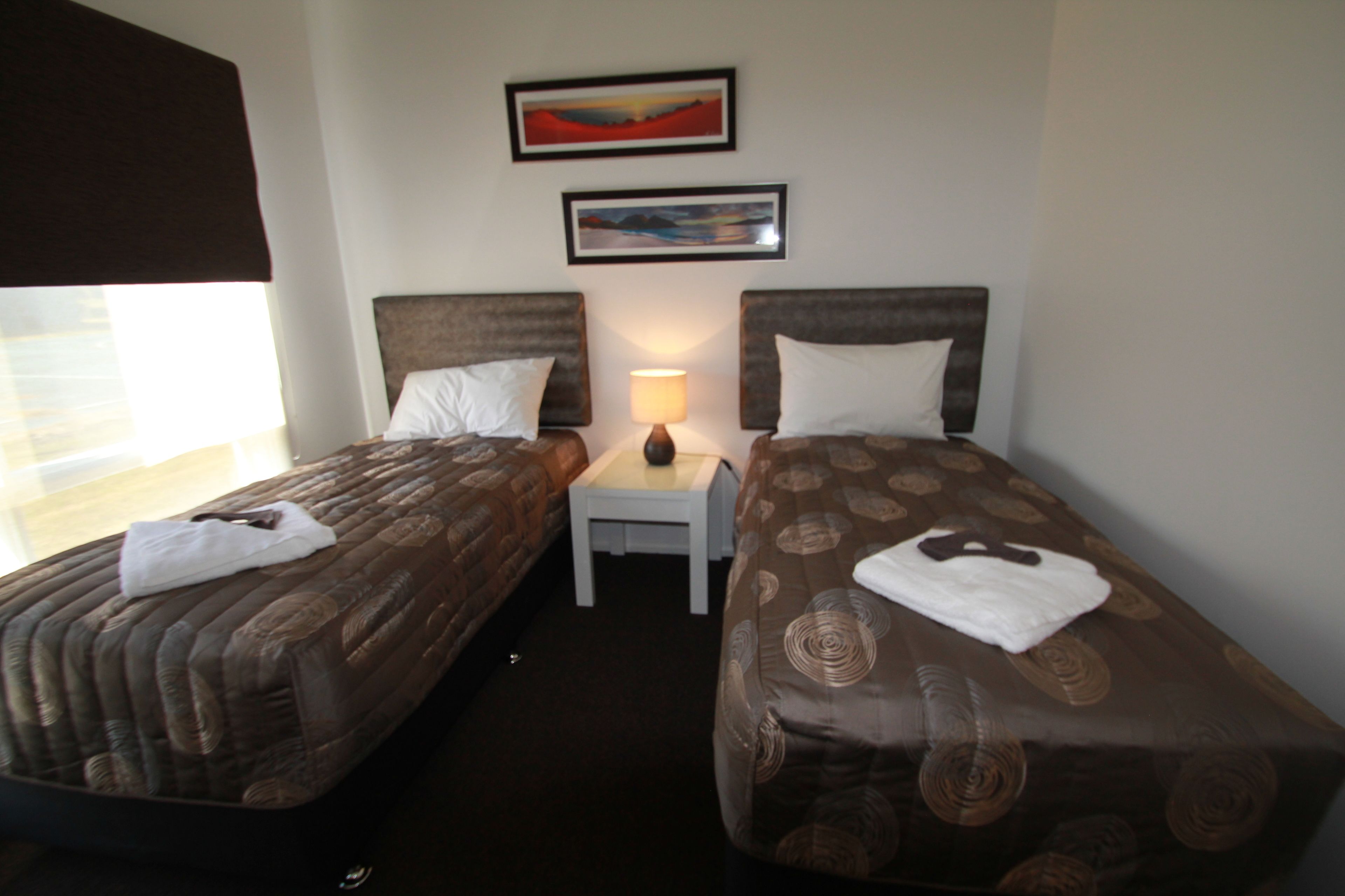 A hotel room with two single beds, a nightstand with a lamp, and two framed pictures on the wall.