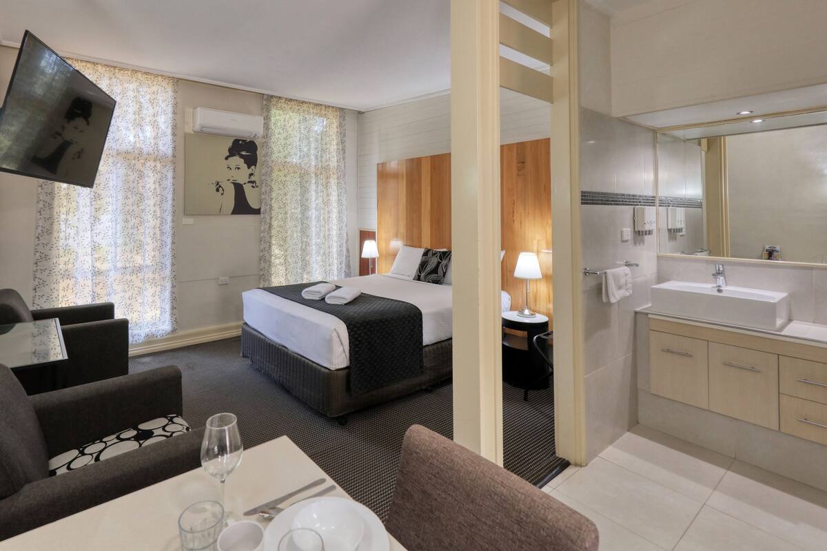 A hotel room with a bed, a desk with a chair, a sofa, a TV, a bathroom with a sink and mirror, and a shower.