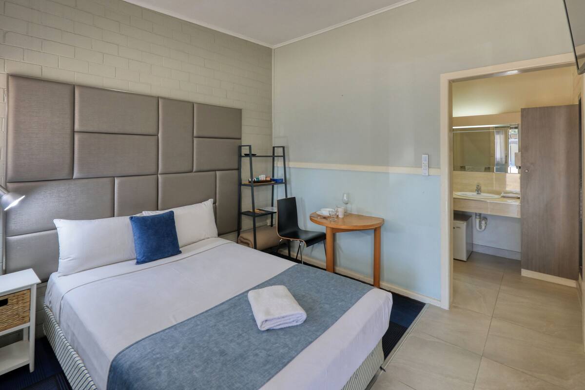 A hotel room with a large bed, a bedside table, a chair, a small round table, and a bathroom visible through an open door.