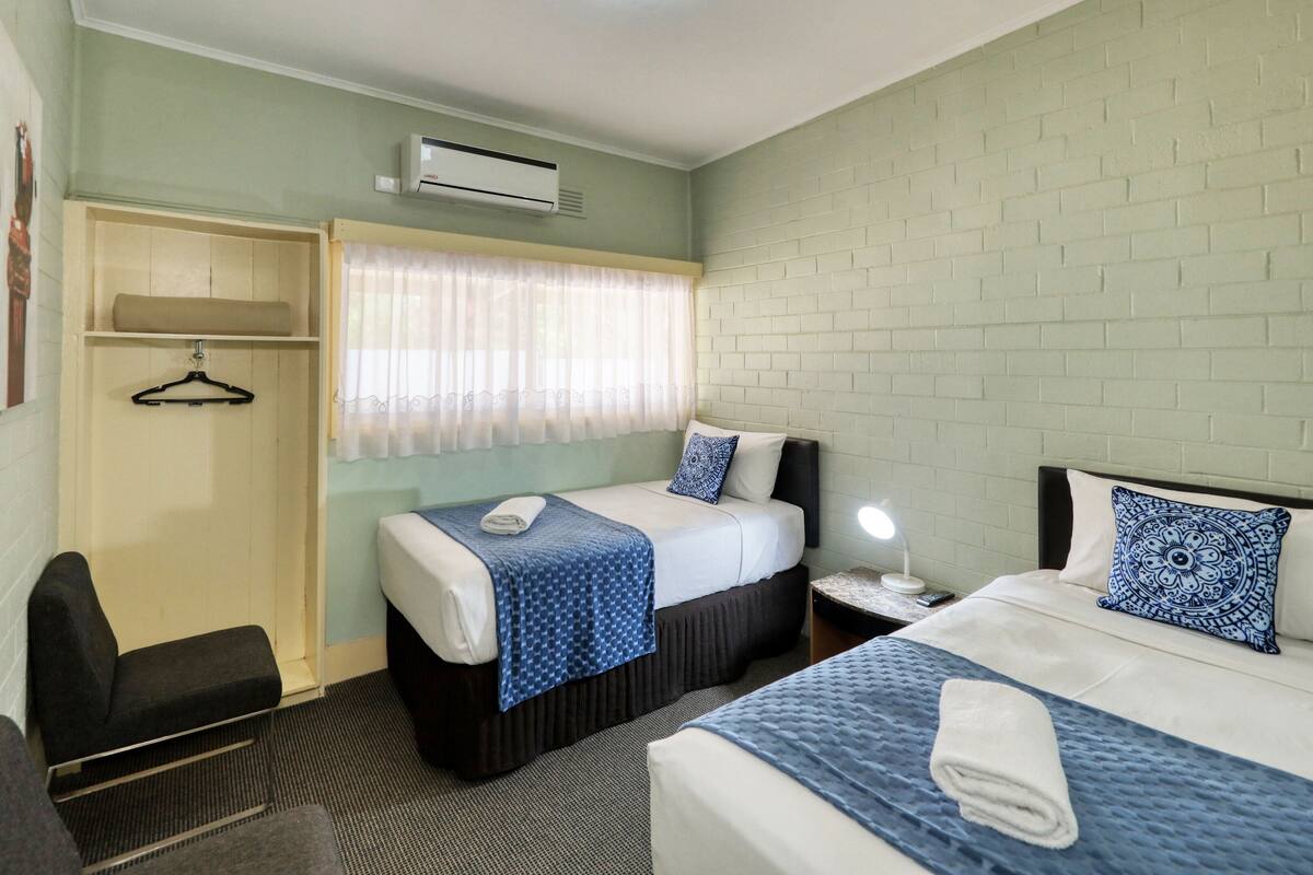 A hotel room with two beds, a bedside table with a lamp, a hanging coat rack, and an air conditioning unit.