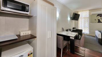 A compact hotel room with a kitchenette, a microwave, a tea sign, a dining table with chairs, and a TV mounted on the wall.