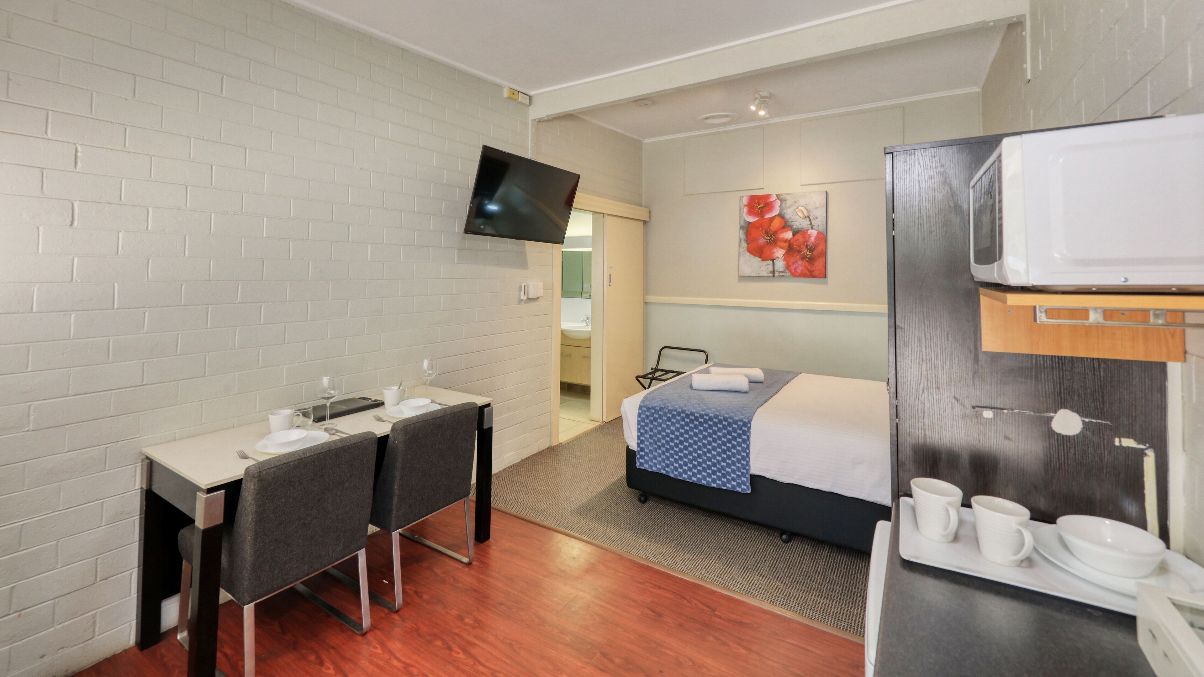 Quarto de hotel com uma cama, uma mesa com duas cadeiras, uma TV fixada na parede e um quadro sobre a cama.