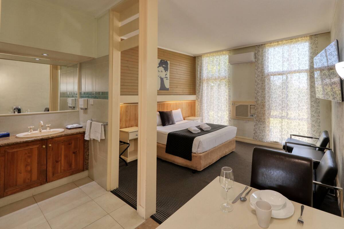 A hotel room with a wooden vanity, a bed with a headboard, a desk with a chair, and a dining table set for two.