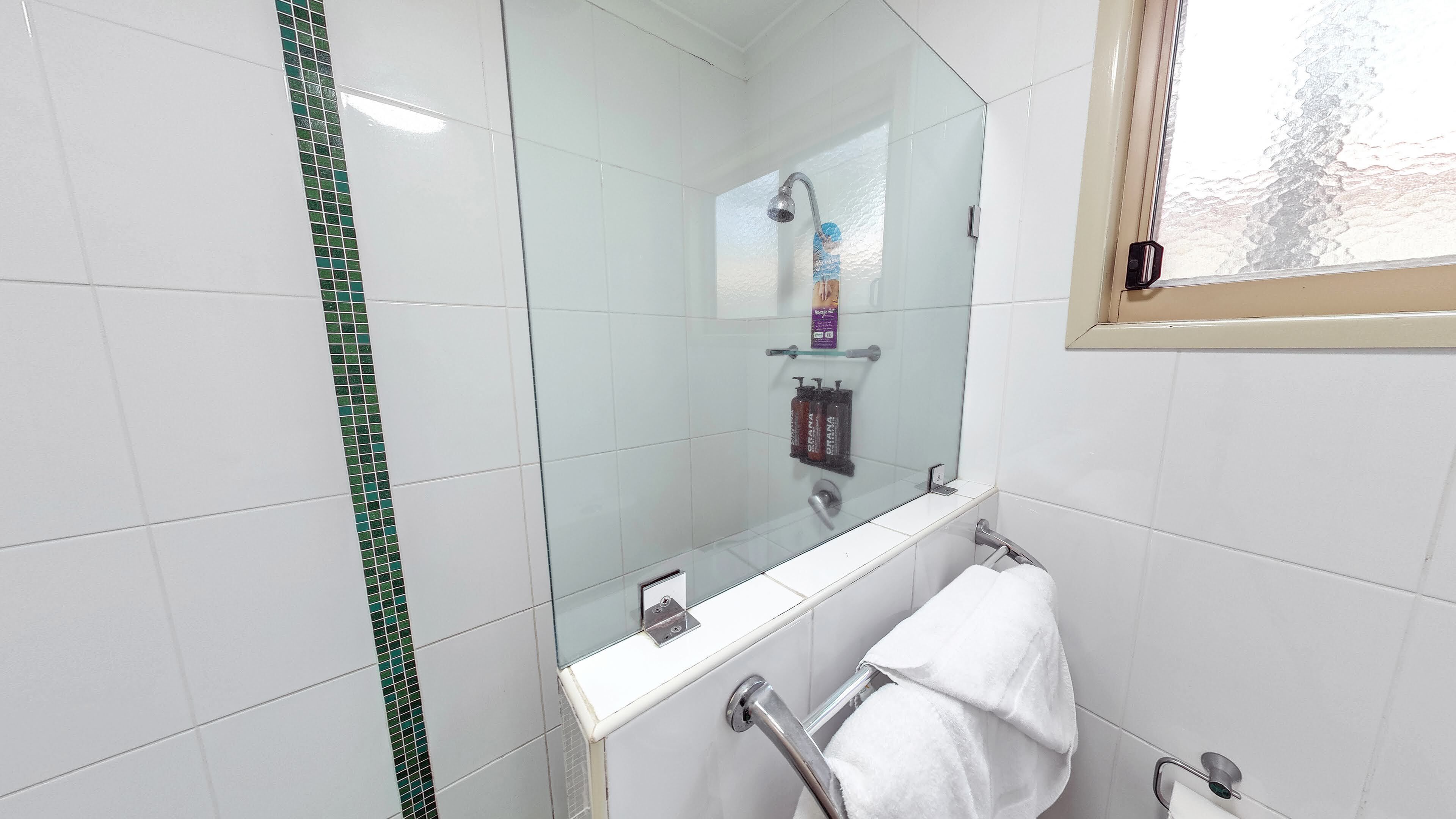 Een moderne badkamer met witte tegels, een douchecabine van glas, een witte handdoekhouder en een raam met uitzicht op bomen.