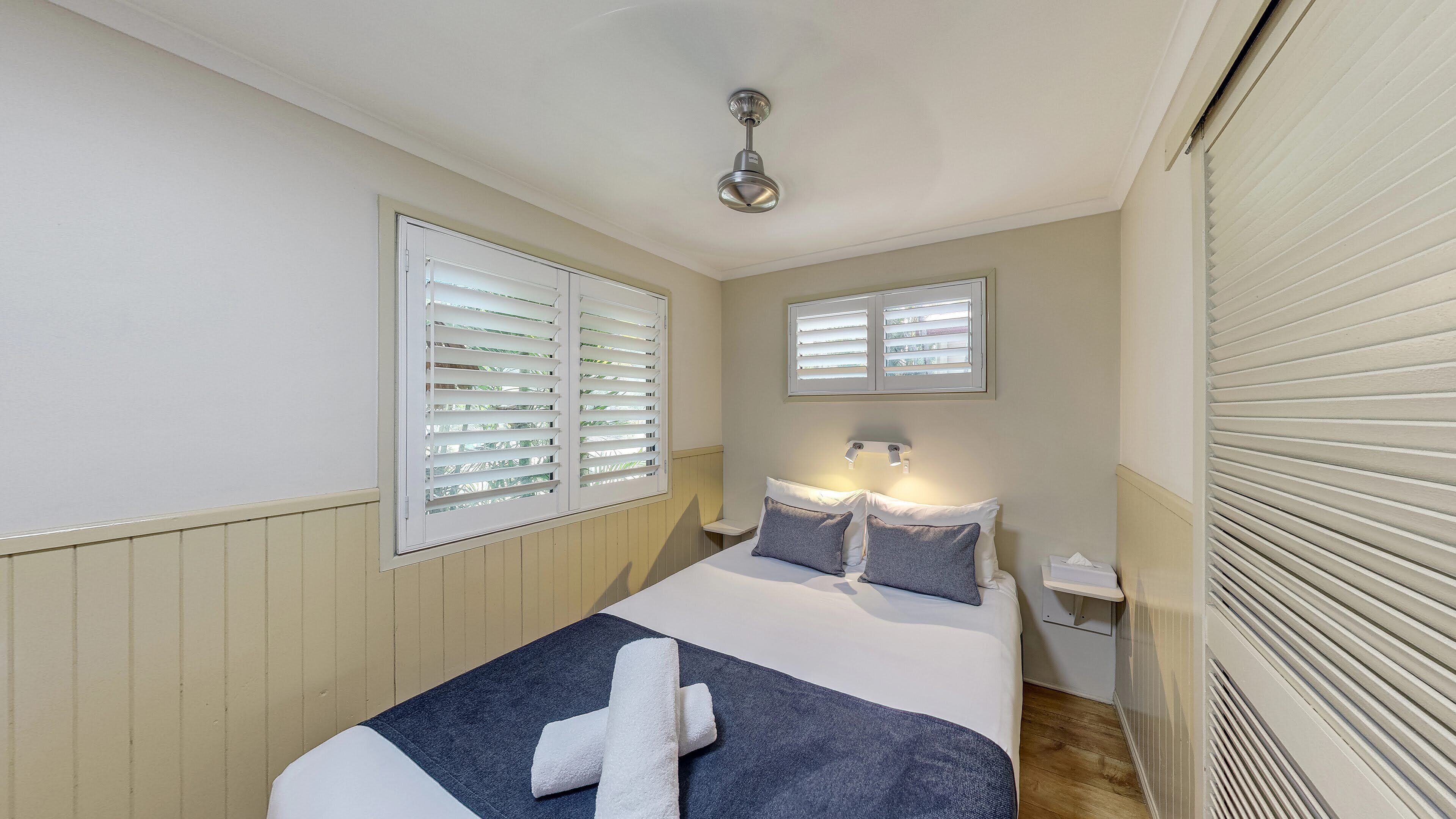 Ein ordentlich bezogenes Bett mit weißer und blauer Bettdecke, zwei Kissen und ein weißes Handtuch. Zwei Fenster mit Jalousien, ein Deckenventilator und ein Nachttisch mit Lampe sind ebenfalls vorhanden.