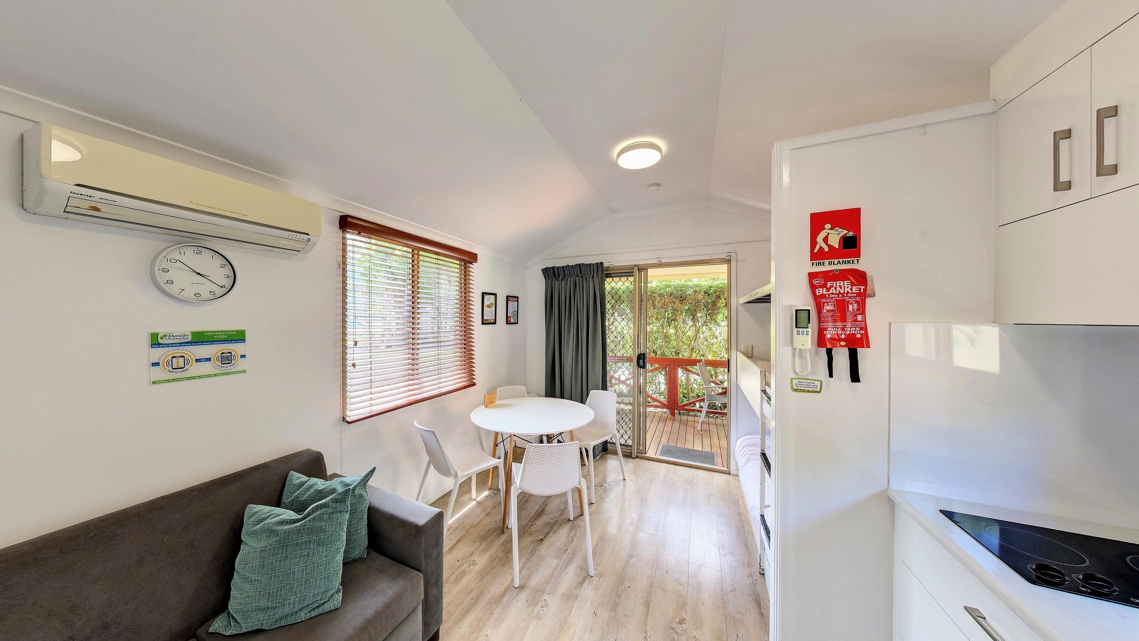 Un espace de vie compact comprenant un coin cuisine, un espace repas avec une table et des chaises, un canapé avec des coussins, une horloge et une couverture anti-feu accrochée au mur.