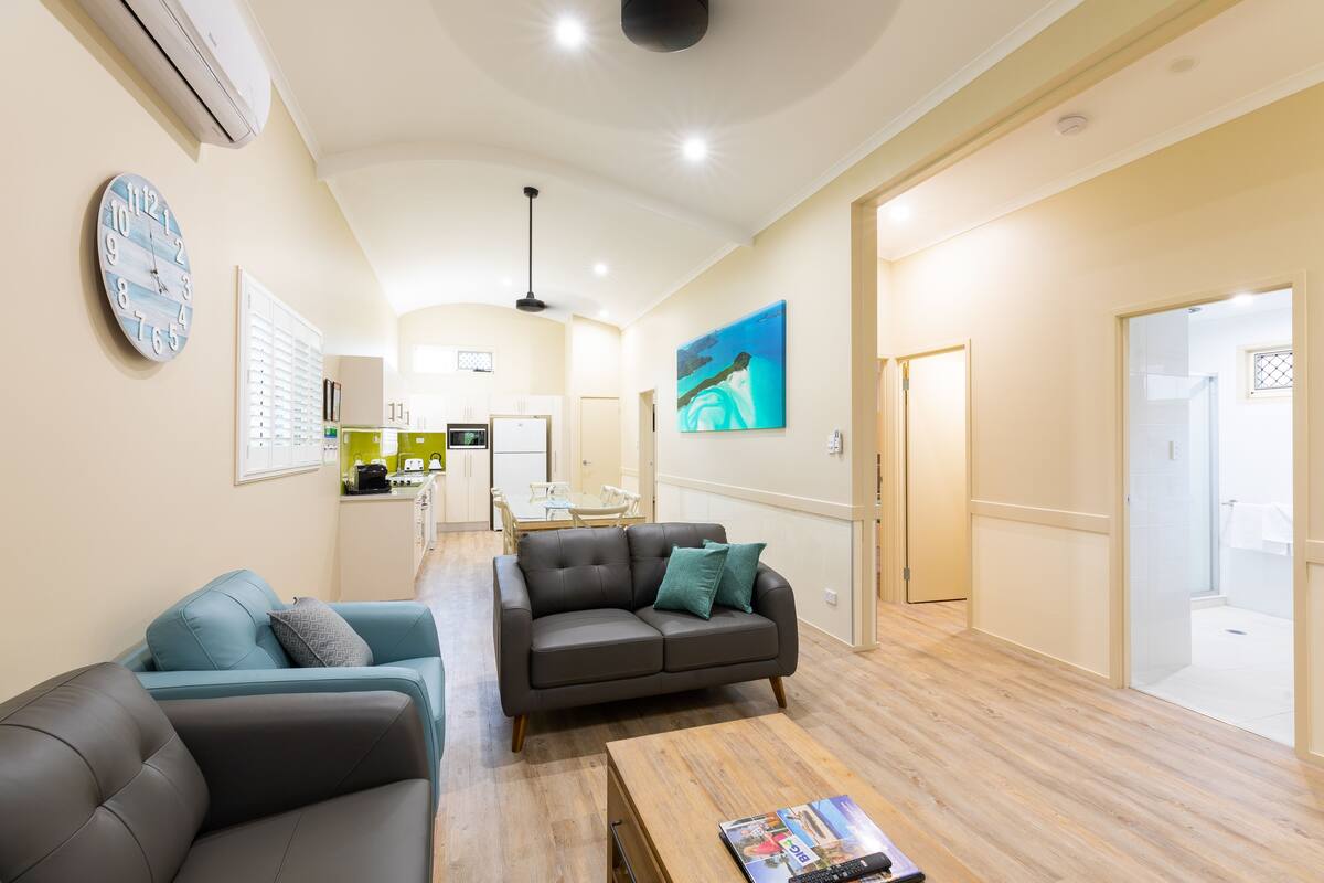 Una sala de estar moderna con un sofá, una mesa de centro y un televisor. Se aprecia una cocina al fondo con una mesa de comedor y sillas.