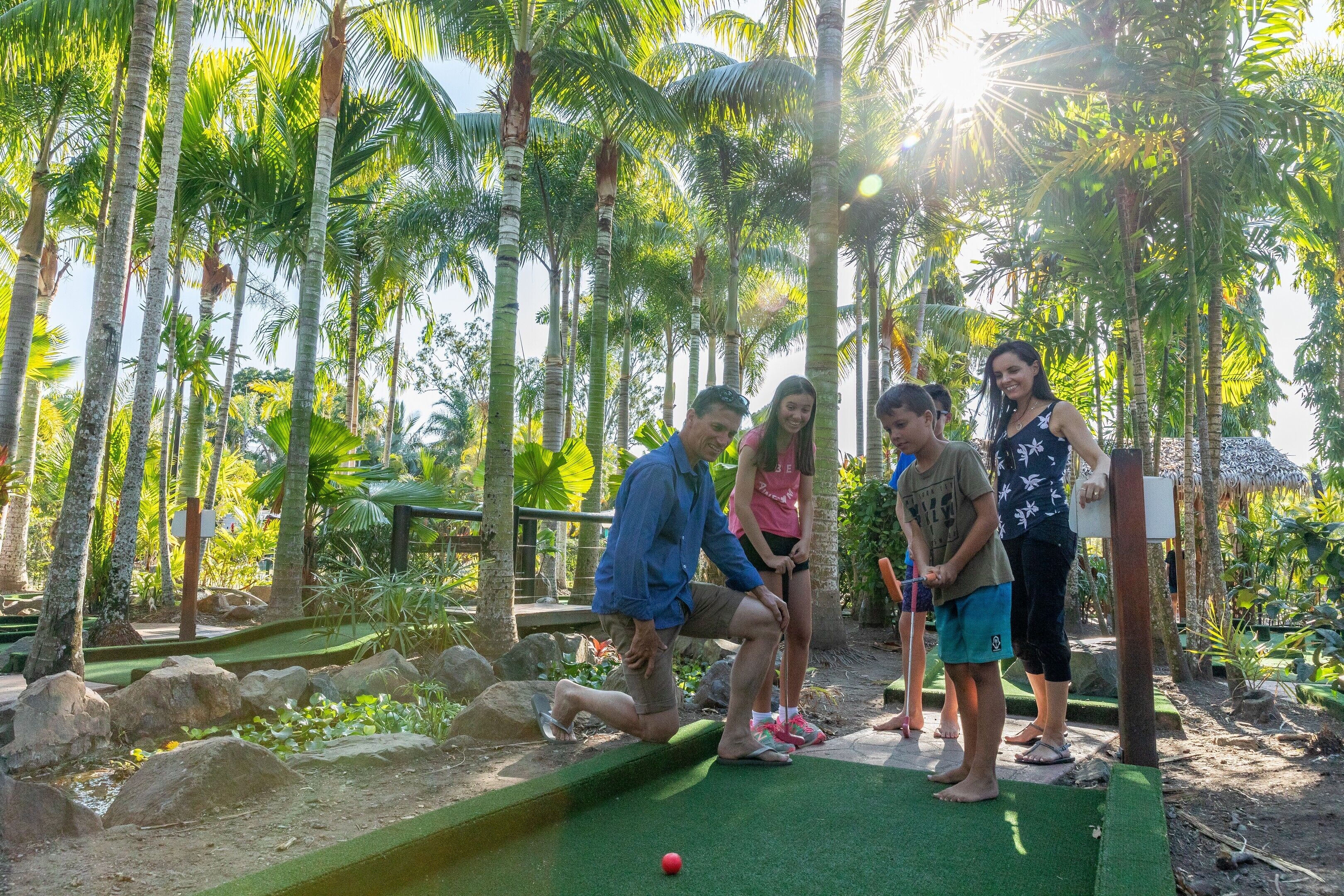 A group of people playing mini-golf in a tropical setting with palm trees and a thatched-roof structure in the background.
