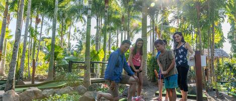 A group of people playing mini-golf in a tropical setting with palm trees and a thatched-roof structure in the background.