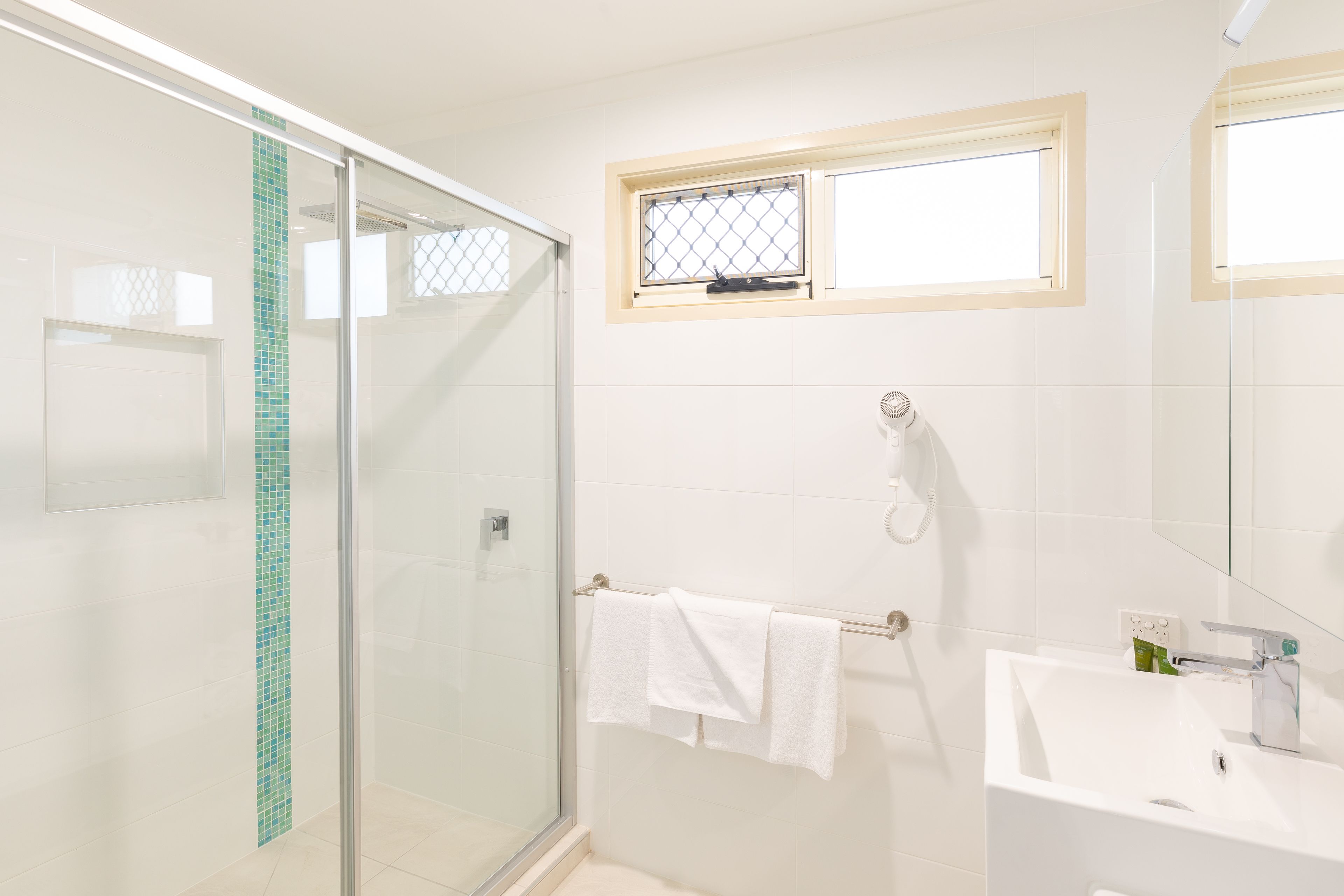 Ein Badezimmer mit Duschkabine aus Glas, einem Fenster mit Metallgitter, einem Handtuchhalter mit zwei weißen Handtüchern und einem weißen Waschbecken mit Spiegel darüber.