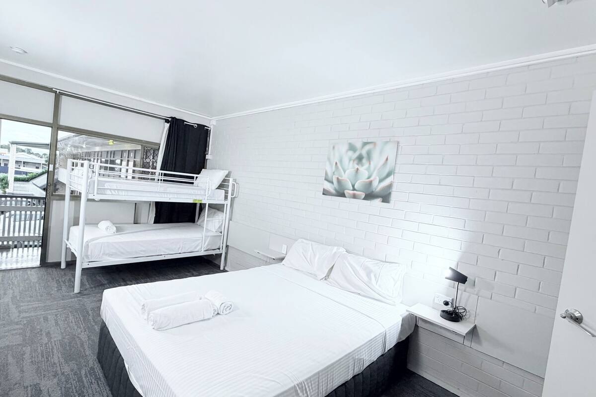 A hotel room with a bunk bed, a single bed, a balcony, and a white brick wall.