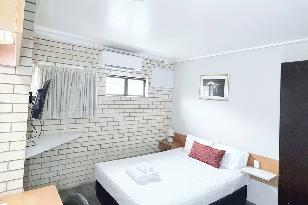 A hotel room with a brick wall, a bed with white bedding and a red pillow, an air conditioning unit, a window with curtains, a framed picture on the wall, and a bedside table with a lamp.