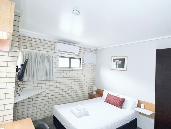 Una habitación de hotel con pared de ladrillo, una cama con ropa blanca y un almohadón rojo, un equipo de aire acondicionado, una ventana con cortinas, un cuadro enmarcado en la pared y una mesita de noche con lámpara.