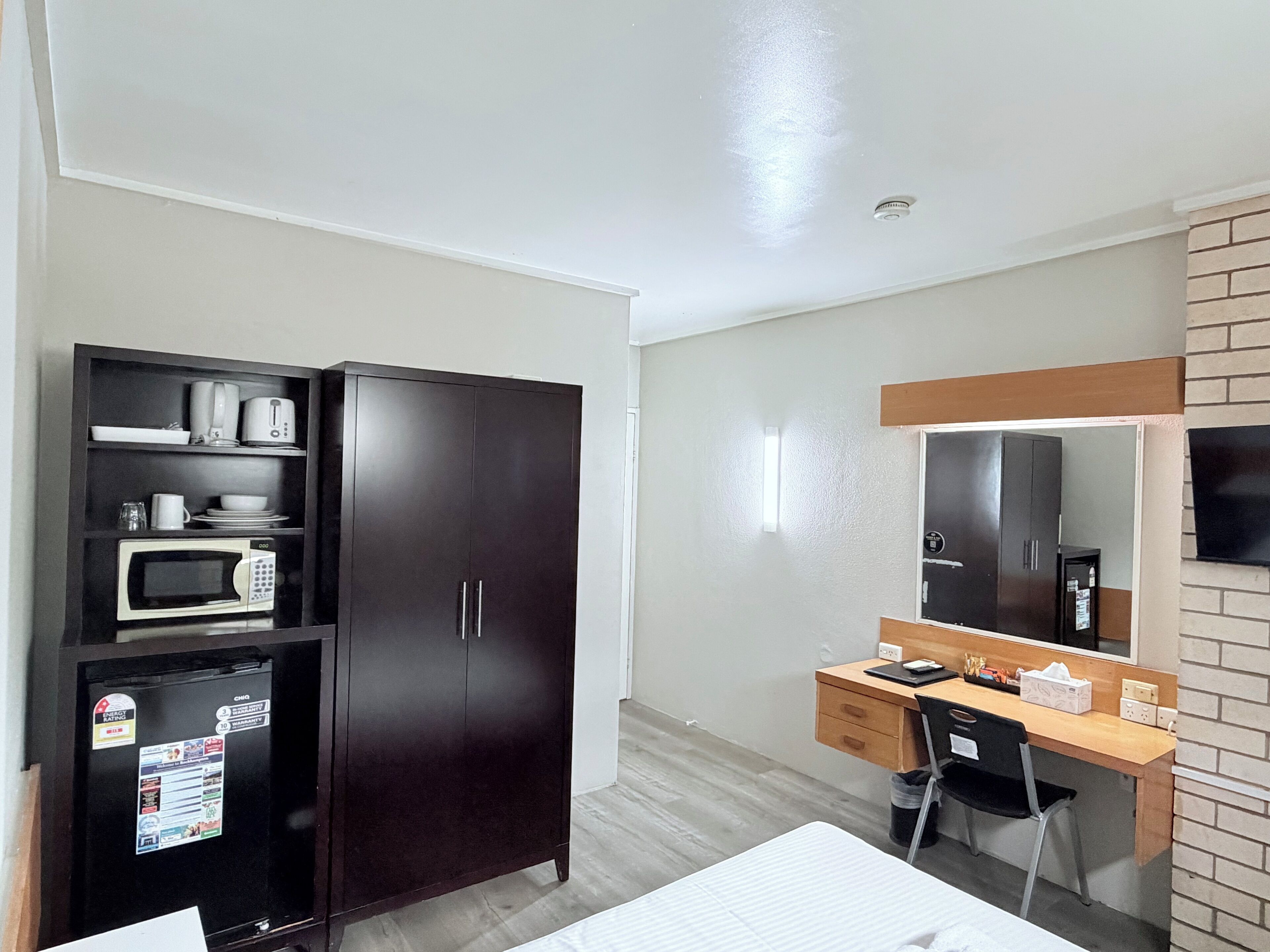 A hotel room with a dark wooden wardrobe, a microwave, a desk with a chair, and a large mirror.