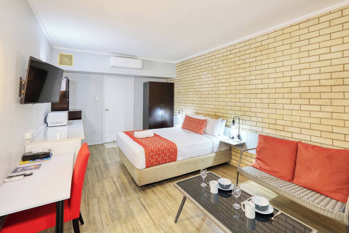 A modern hotel room with a bed, a desk, a TV, and a brick wall.