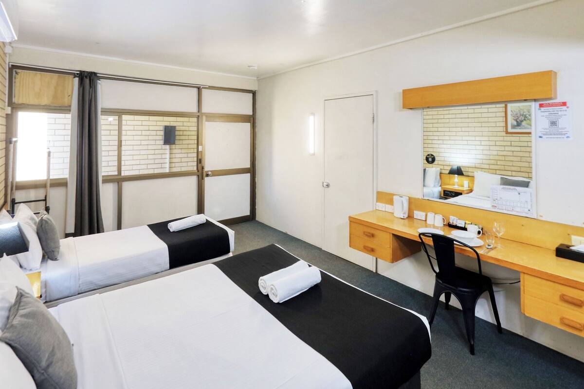 A hotel room with two beds, a desk, a chair, a mirror, and a window with blinds.