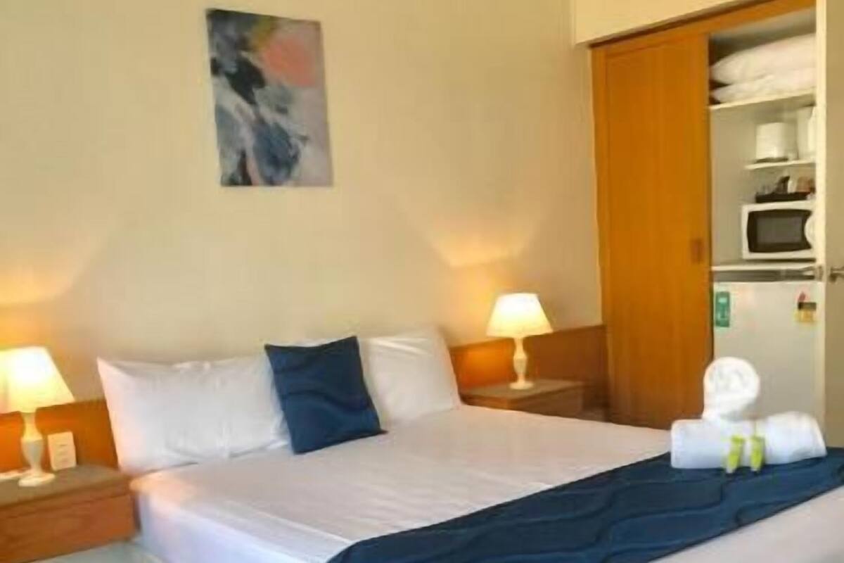 A hotel room with a bed, two bedside lamps, a painting on the wall, and a wardrobe with folded towels.