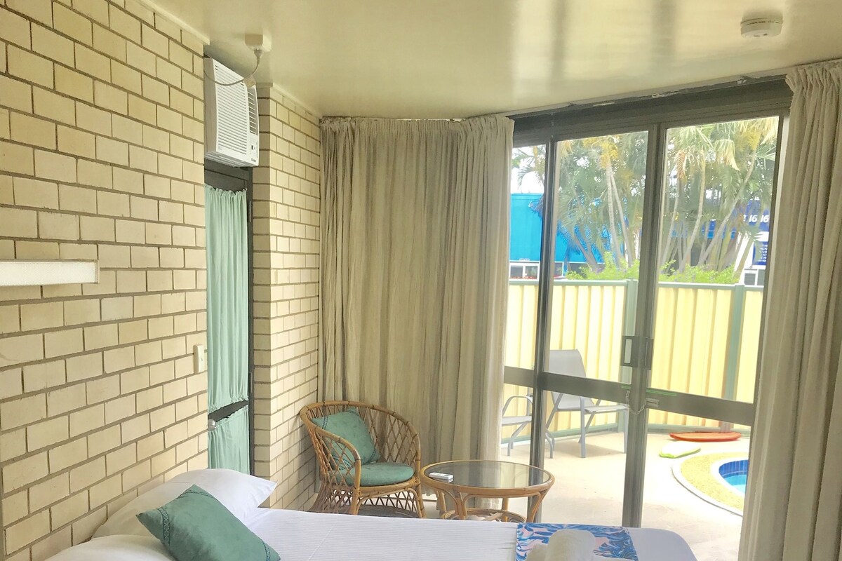 A hotel room with a large bed, a wicker chair, a small table, and a view of a pool.