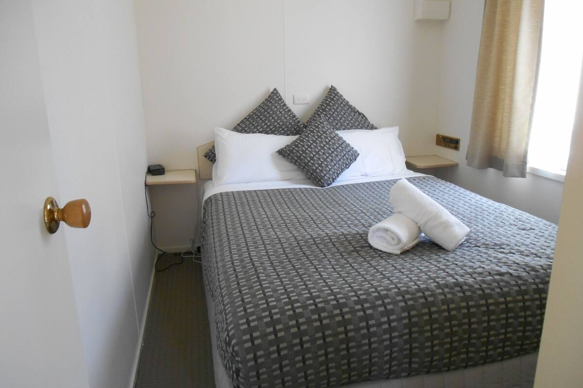A neatly made bed with a checkered bedspread and two pillows, a bedside table with a phone, and a door with a wooden knob.