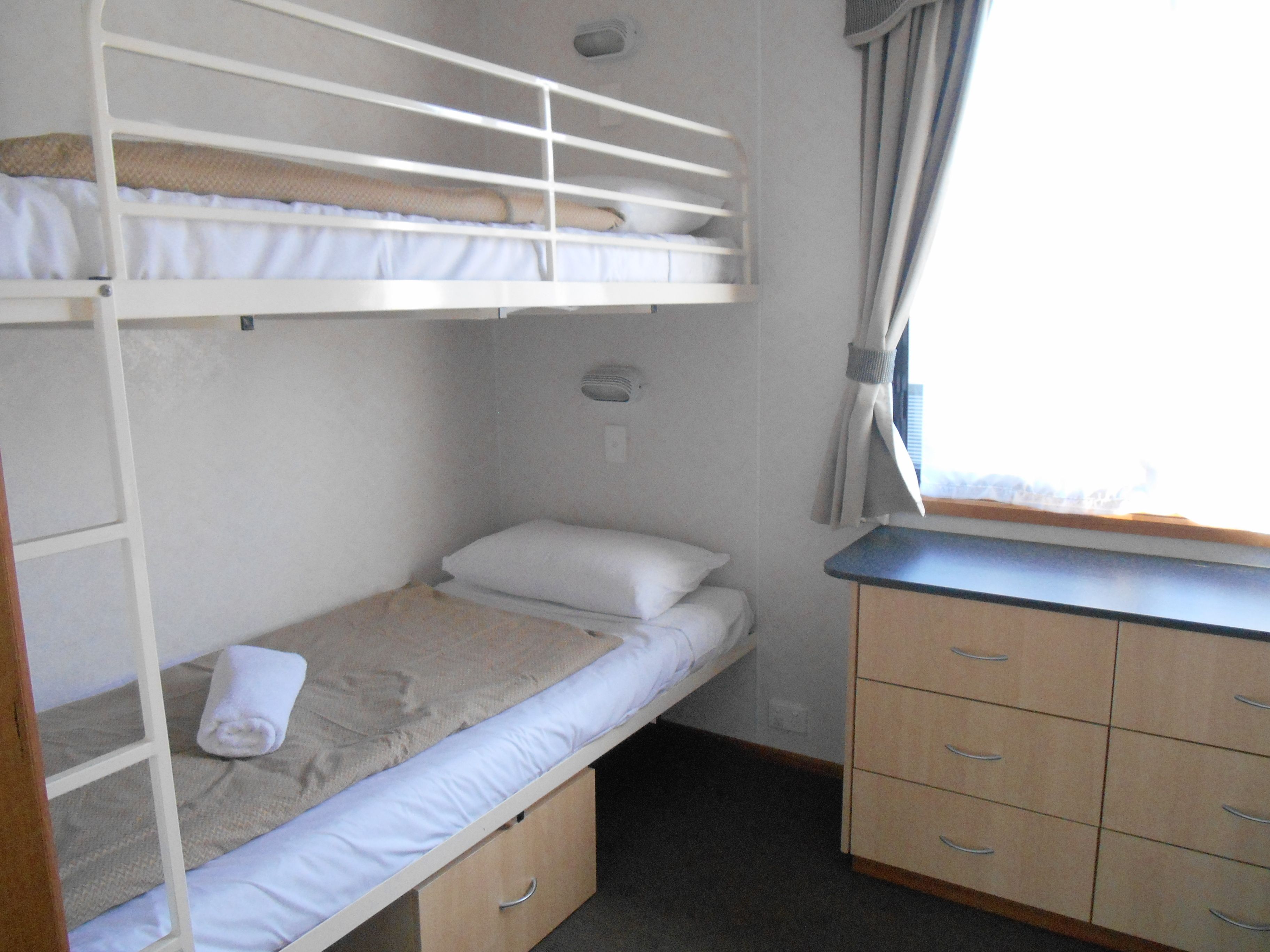 Ein Stockbett mit unterem Bett, einem Einzelbett, einer hölzernen Kommode mit Schubladen, einem kleinen Wandheizgerät und einem Fenster mit Vorhängen.