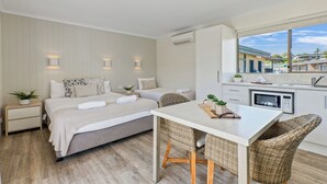 A modern bedroom with a large bed, bedside tables, a dining area with wicker chairs, and a view of the outside through a window. - Tasman Holiday Parks – Merimbula (Merimbula)