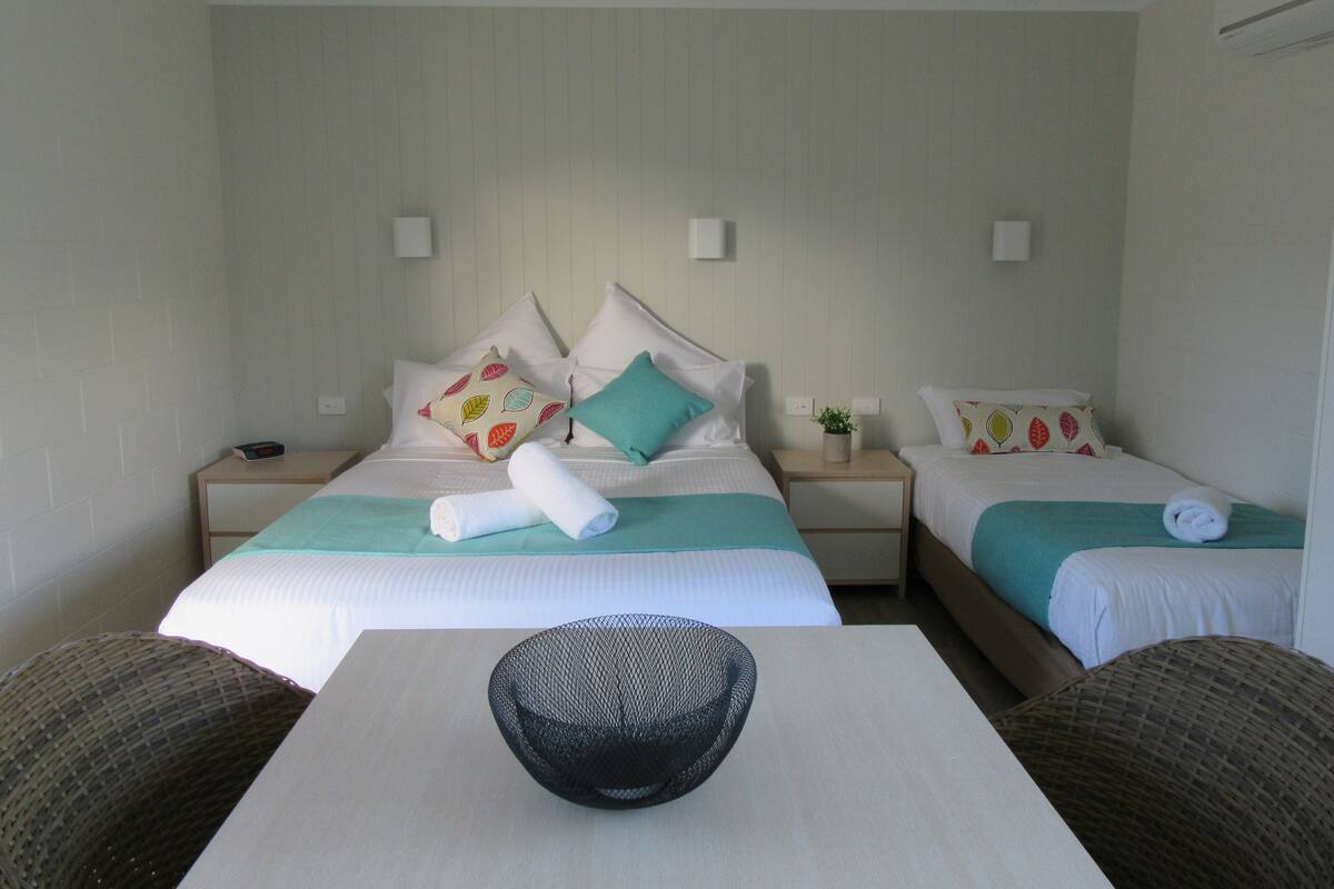 A hotel room with two beds, each with a pillow and a rolled towel. There is a small table with a bowl in the center of the room.