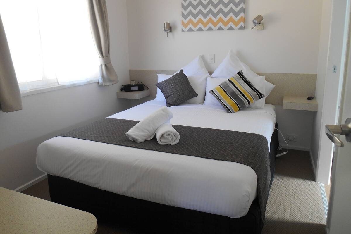 A neatly made bed with white and gray bedding, a striped pillow, and a rolled towel on a dark headboard.