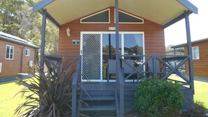 A wooden cabin with a covered porch, a number sign, and a window with a metal screen. - Tasman Holiday Parks – Merimbula (Merimbula)